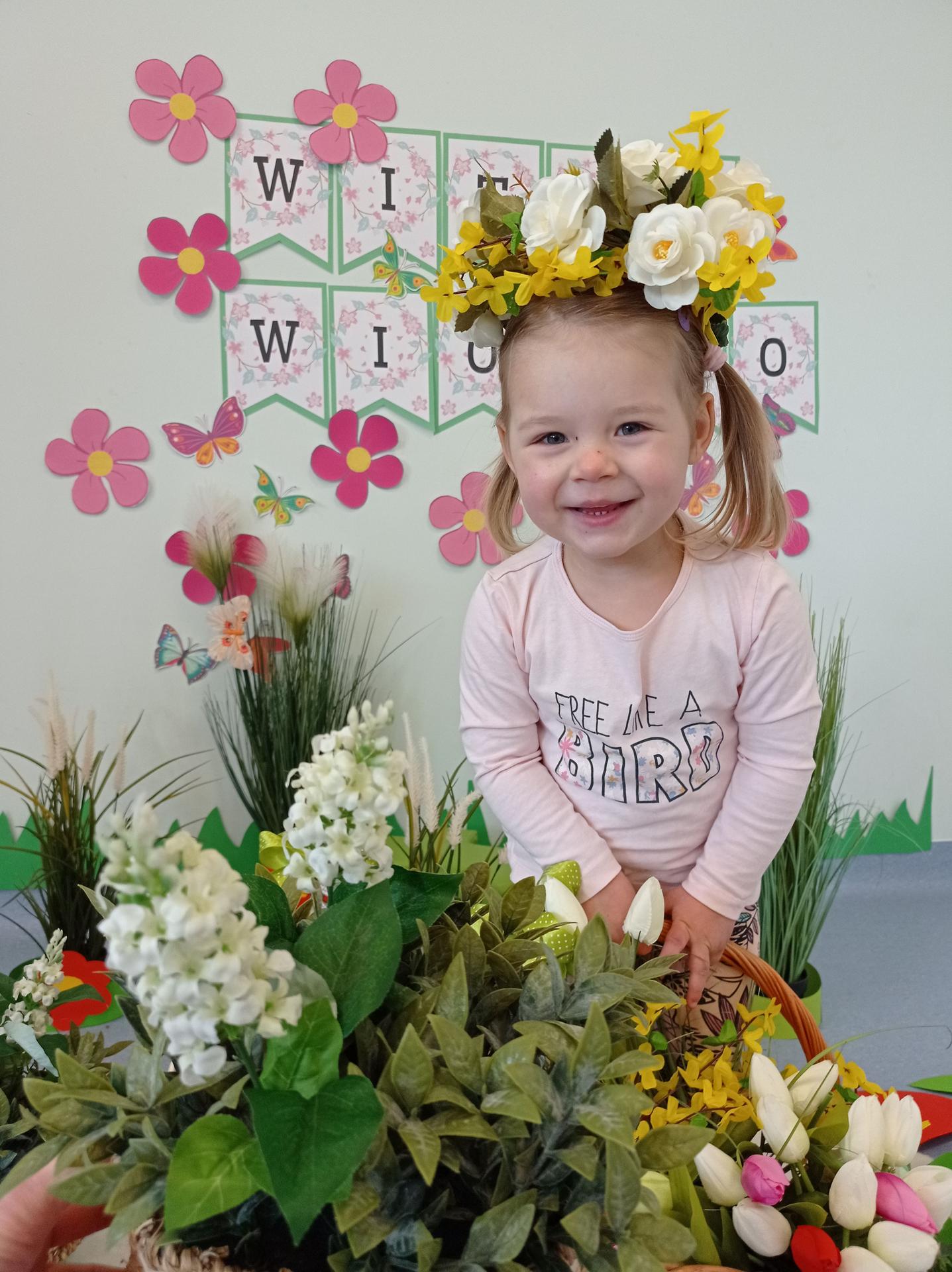 Na zdjęciu widać dziewczynkę ok 3 lat z wiankiem z kwiató na głowie. Dziewczynka jest uśmiechnięta. Za dziewczynką widać napis WITAJ WIOSNO. Dziewczynka stoi wśród kwiatów.