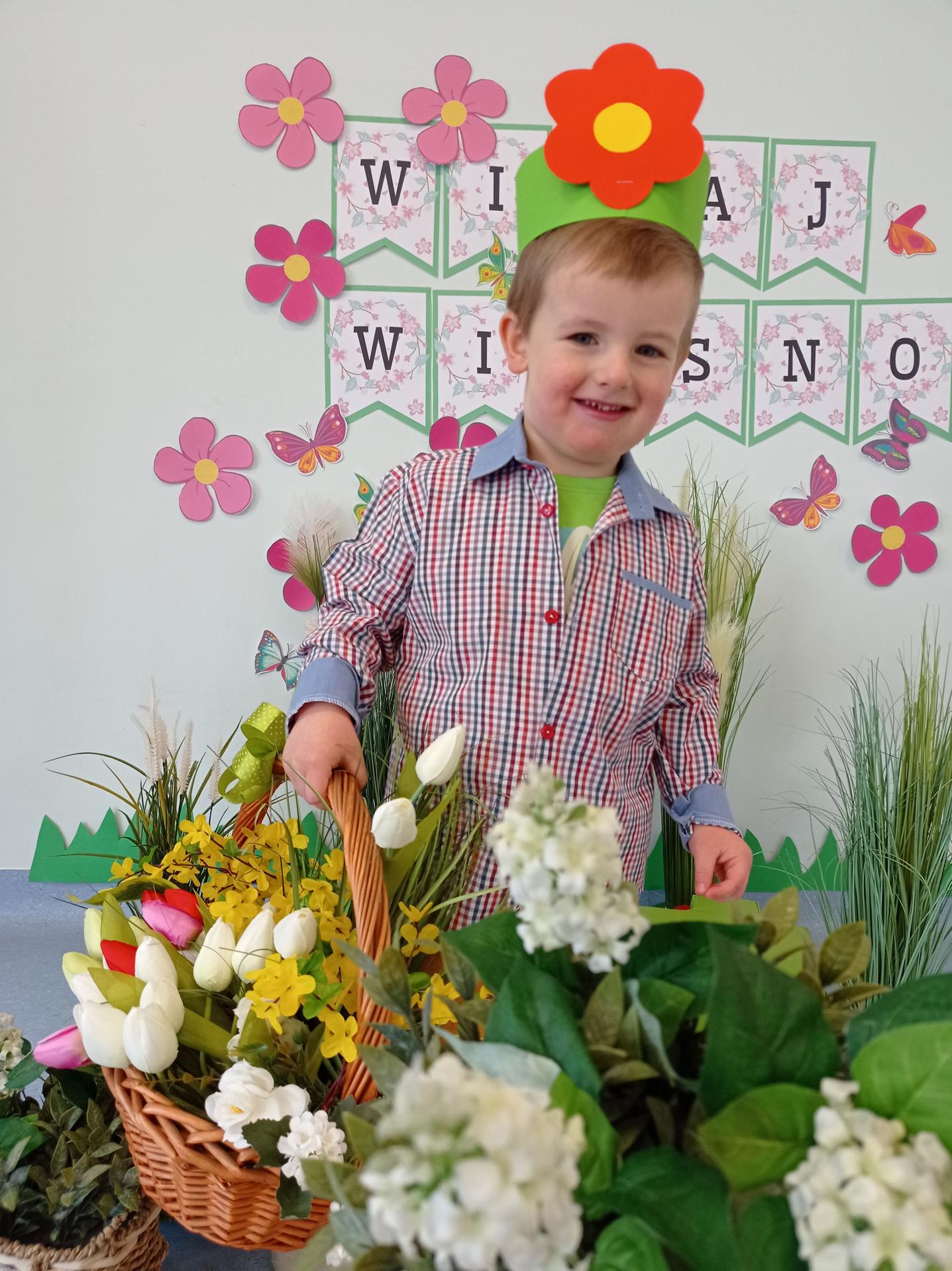 Na zdjęciu widać chłopca w wieku ok 3 lat. Chłopiec jest uśmiechnięty. Na głowie ma opaskę z kwiatkiem. W ręku trzyma kosz z kwiatami. Za nim widać napis WITAJ WIOSNO.